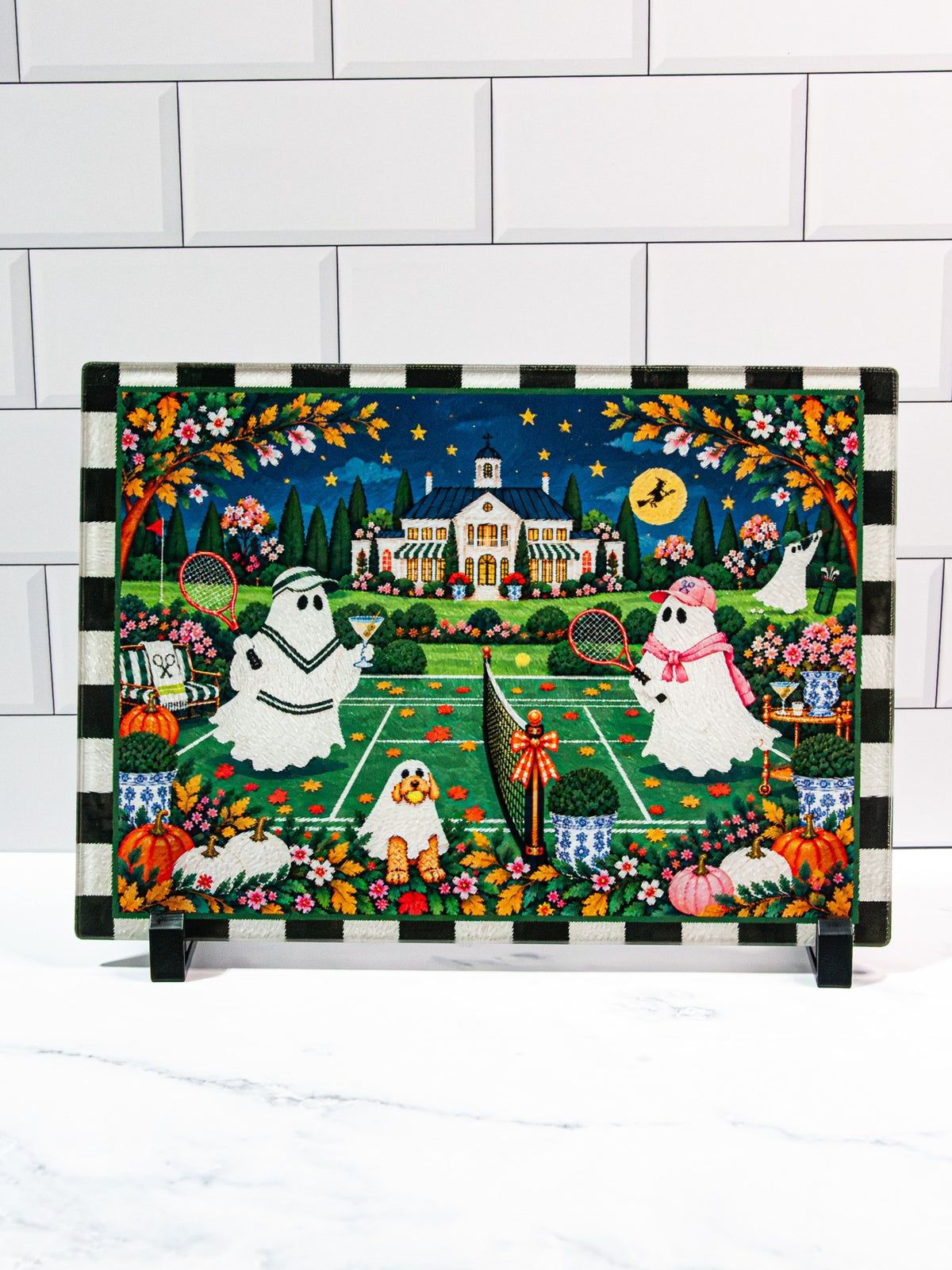 BOOugie Night Halloween glass cutting board with tennis-playing ghosts, a moonlit clubhouse, pumpkins, flowers, and a black-and-cream checked border.