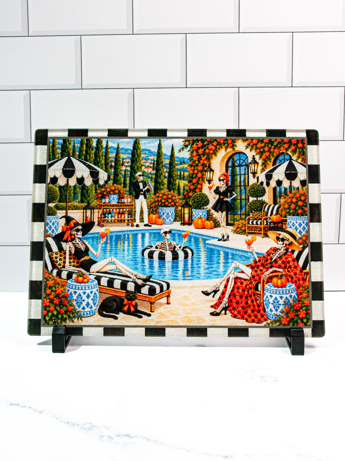 The Bone Vivants Halloween glass cutting board featuring fashionable skeletons enjoying cocktails beside a swimming pool, pumpkins, blue-and-white planters, and a black-and-cream checked border.