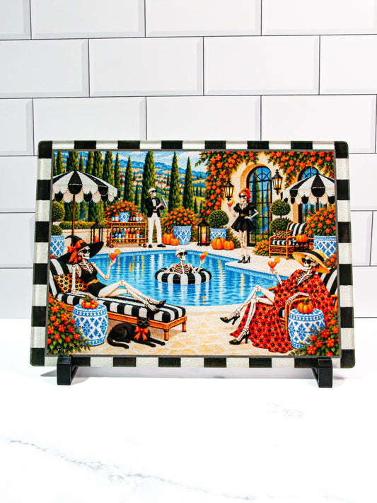 The Bone Vivants Halloween glass cutting board featuring fashionable skeletons enjoying cocktails beside a swimming pool, pumpkins, blue-and-white planters, and a black-and-cream checked border.