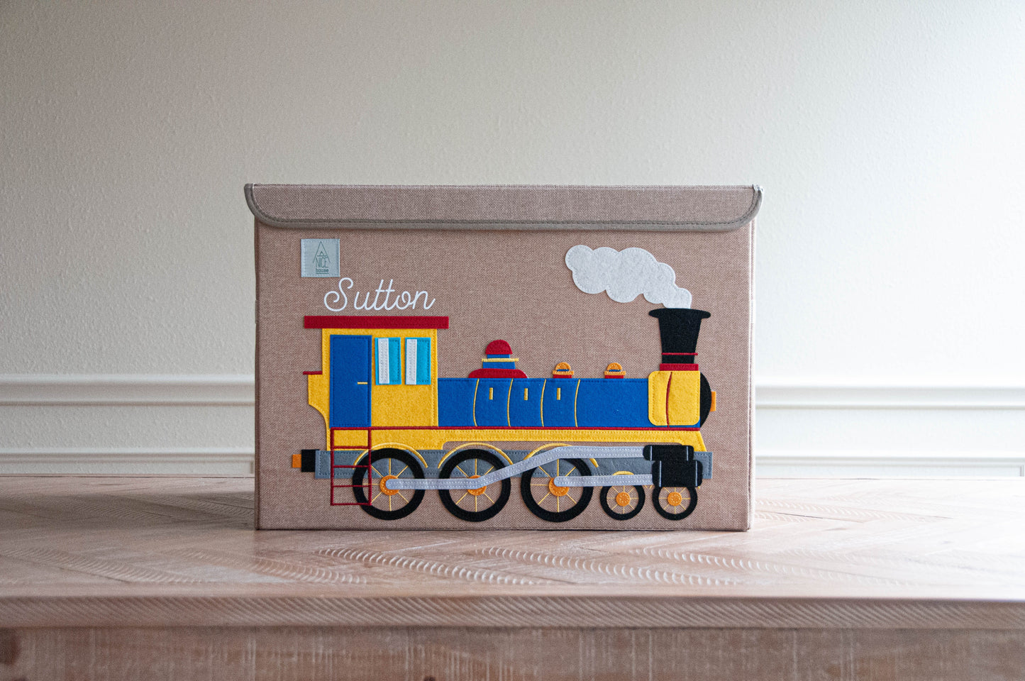 Fabric covered storage bin with lid that is also a personalized toy box. These personalized toy boxes are from A Nice House. This large toy box features a train appliquéd design on the front.